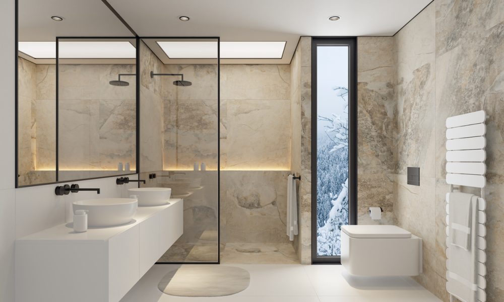 A sleek and sophisticated modern bathroom featuring large limestone or travertine tiles on the walls and floors. The space includes two elegant sinks, a minimalist toilet, and a spacious shower with a contemporary shower head. The expansive mirror enhances the sense of openness, while the natural stone elements provide a serene and luxurious atmosphere. This image is perfect for interior design projects, bathroom renovations, or showcasing high-end bathroom furnishings and fixtures.